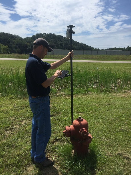 A person is conducting an inspection of a red fire hydrant using a tablet and a surveying pole. 