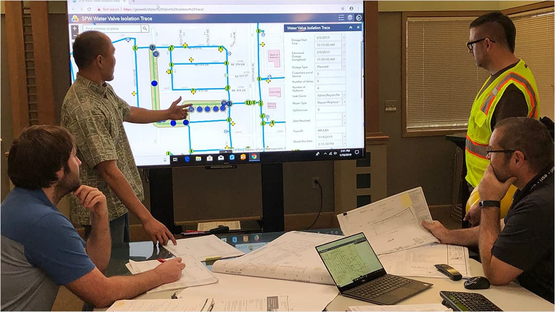 A group of professionals gathers around a large screen displaying a detailed water system map.