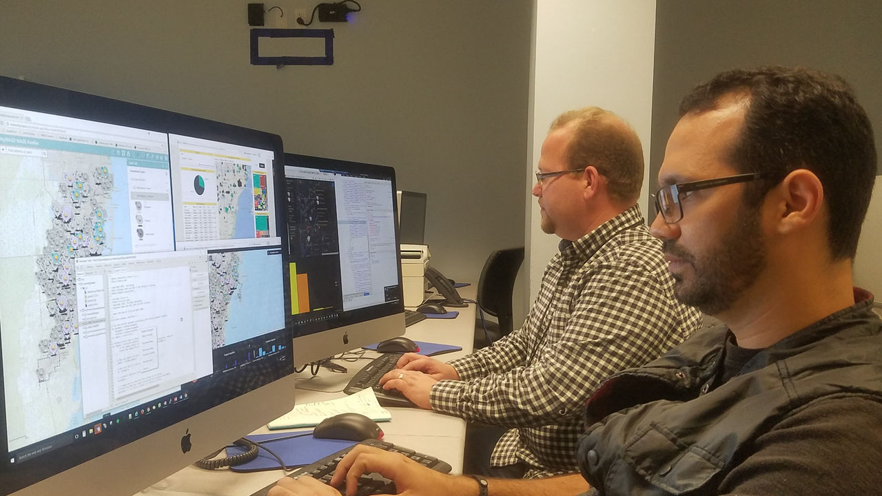 Two individuals are working at a desk, focusing on multiple desktop monitors displaying data visualizations and maps.