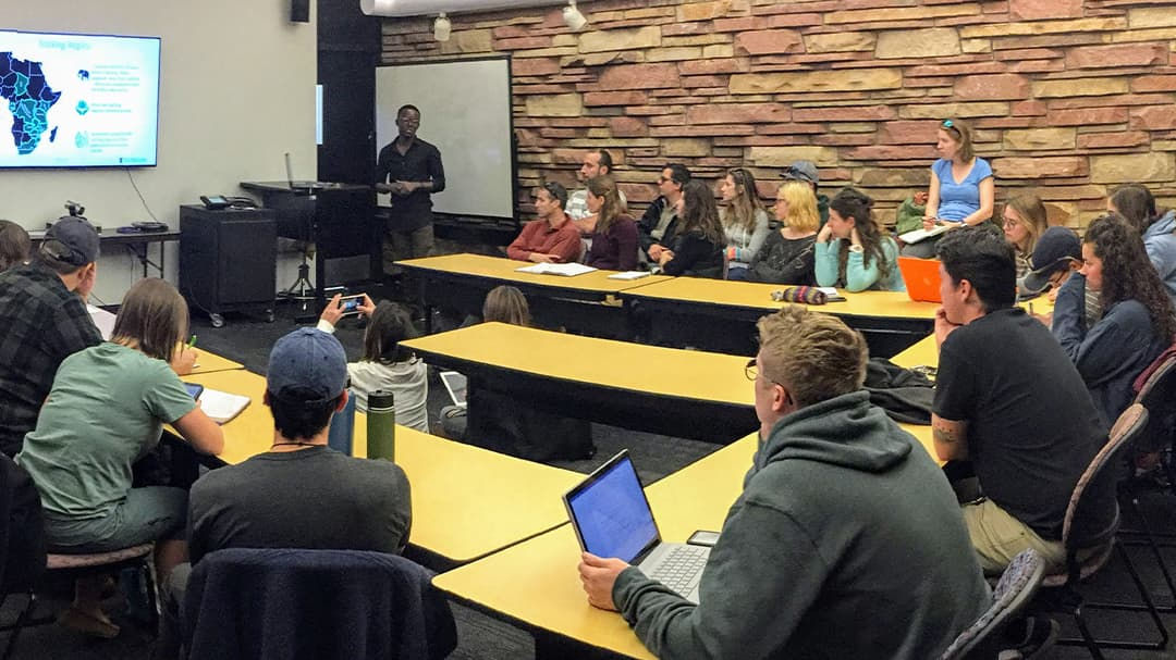 A speaker presents to a group of students in a classroom setting with a stone accent wall. 
