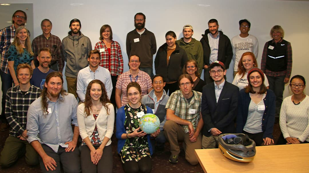 A diverse group of people gathered indoors for a group photo. One individual holds a globe, while a helmet is placed on a table in the foreground. 