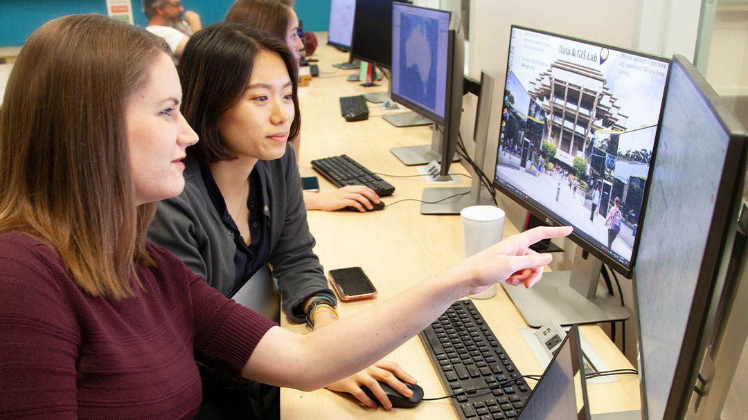 Two women are seated at a computer workstation in a modern office setting, collaborating on a project.