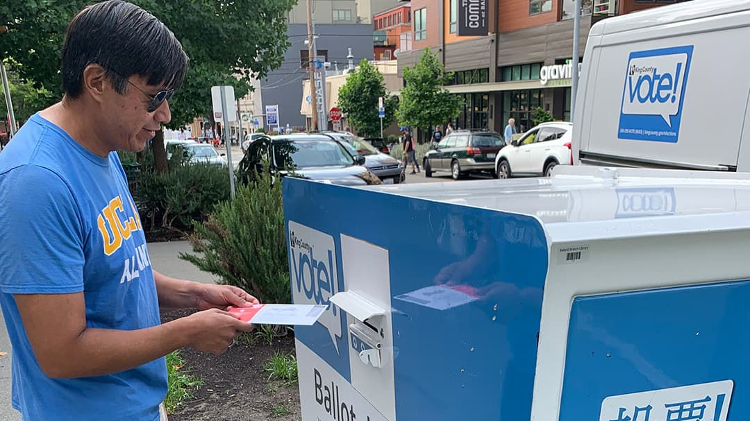 A man wearing a blue shirt is placing a ballot into a voting drop box in an urban setting. 