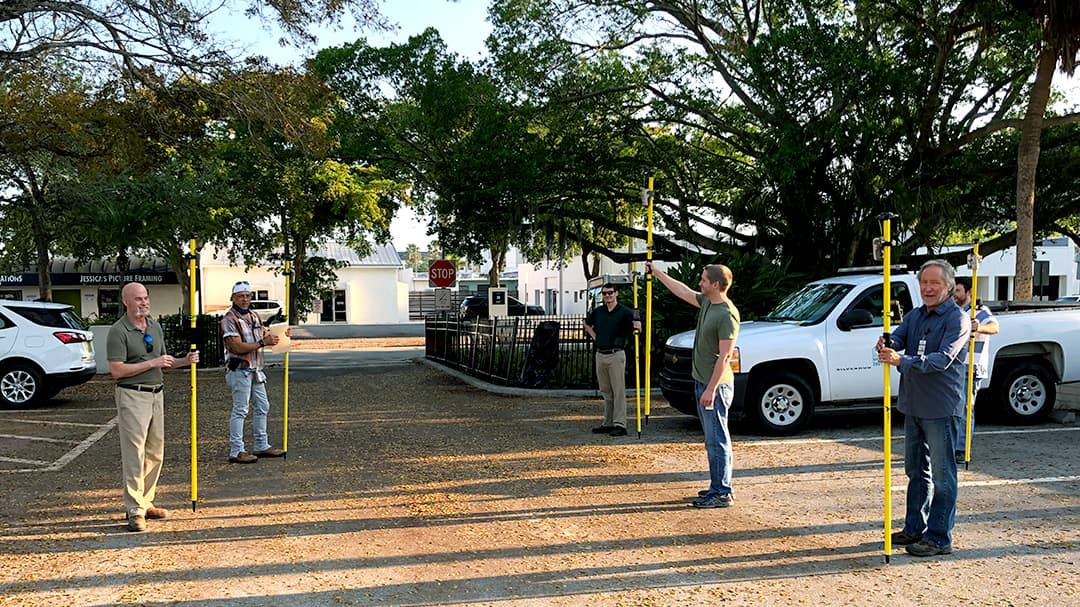 A group of five adults holding yellow measuring poles in a parking lot surrounded by trees and vehicles.