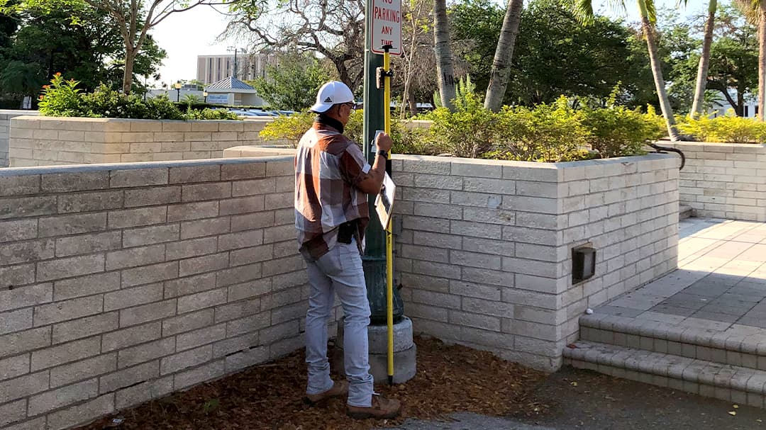 A male surveyor wearing a white hard hat and plaid shirt operates a yellow measuring tool near a landscaped area with brick walls. 
