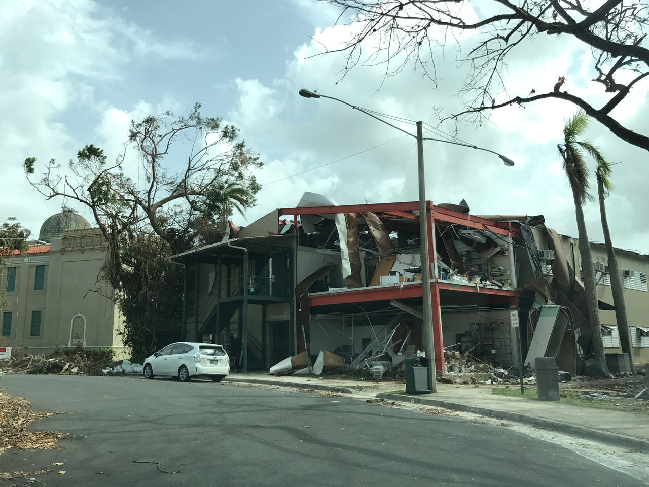 A partially collapsed building is shown in an urban area, with debris scattered around. 