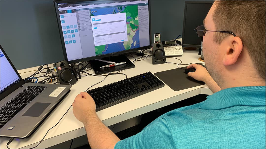 A man in a teal shirt is seated at a desk, using a desktop computer with mapping software displayed on the screen. 