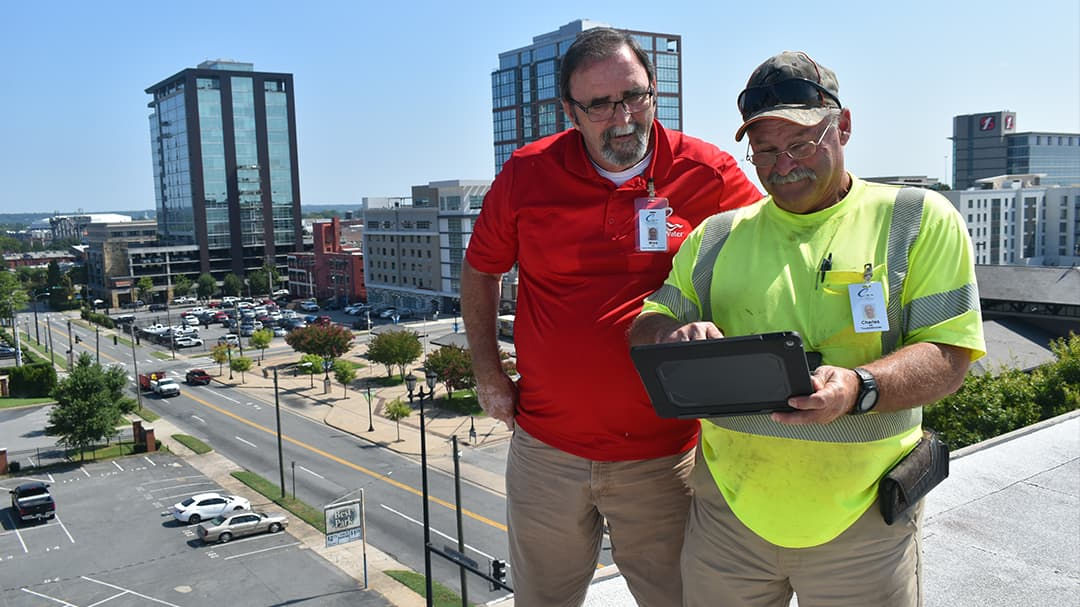 Two workers stand on a rooftop overlooking a cityscape, one holding a tablet device.
