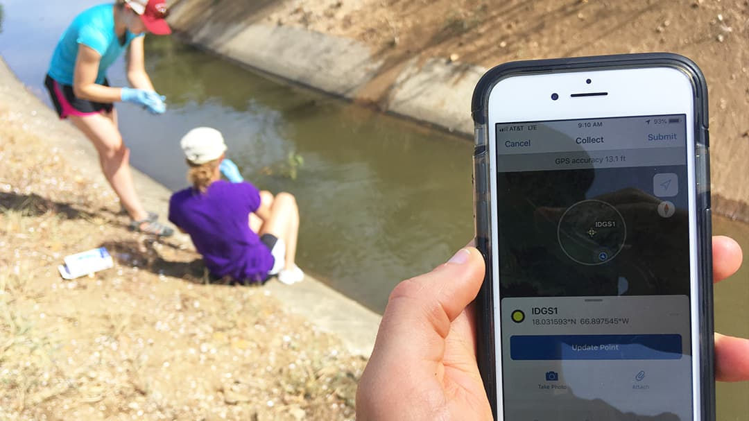 A person holds a smartphone displaying a water sampling app interface while two individuals collect water samples near a canal.