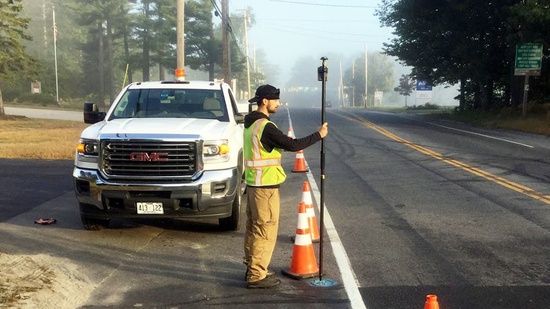 A male surveyor wearing a safety vest and cap is seen working on the roadside with a surveying pole. 