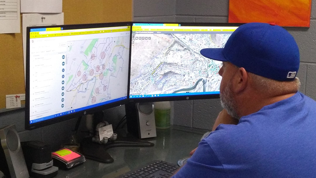 A man wearing a blue cap is seated at a desk, closely examining maps displayed on two computer monitors.