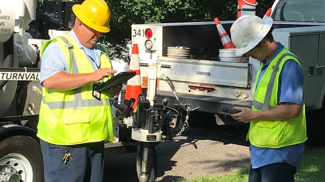 Two utility workers wearing safety gear are inspecting equipment near a utility truck in an outdoor setting. 
