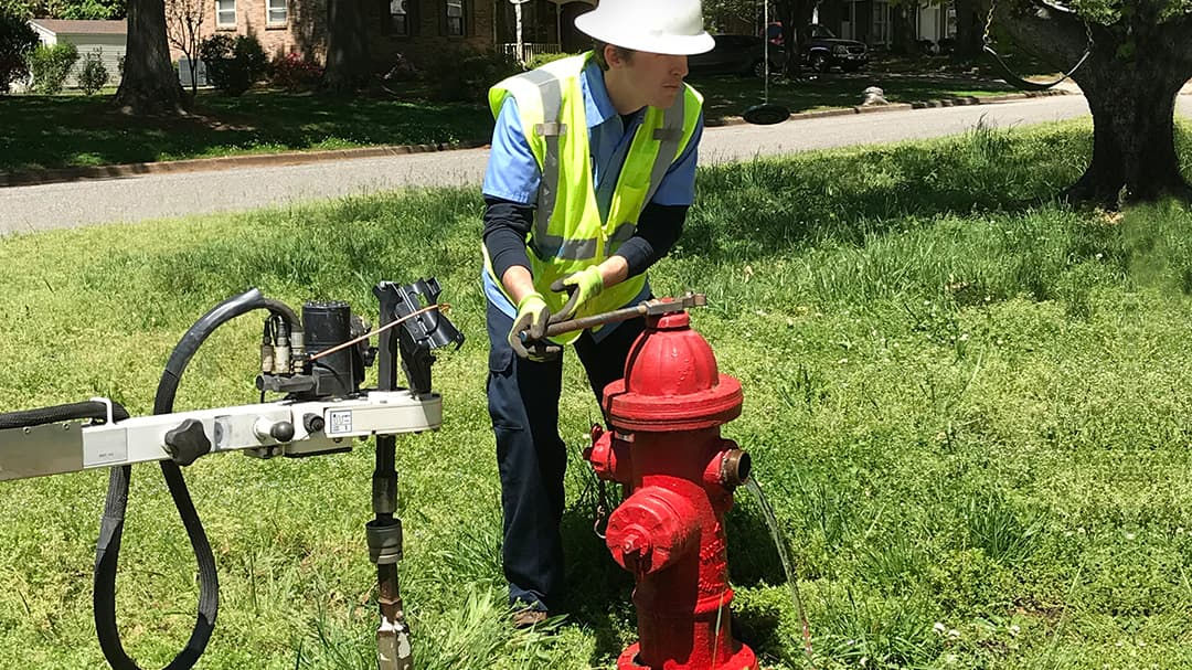 A worker wearing a high-visibility vest and gloves is inspecting a red fire hydrant in a grassy outdoor setting. 