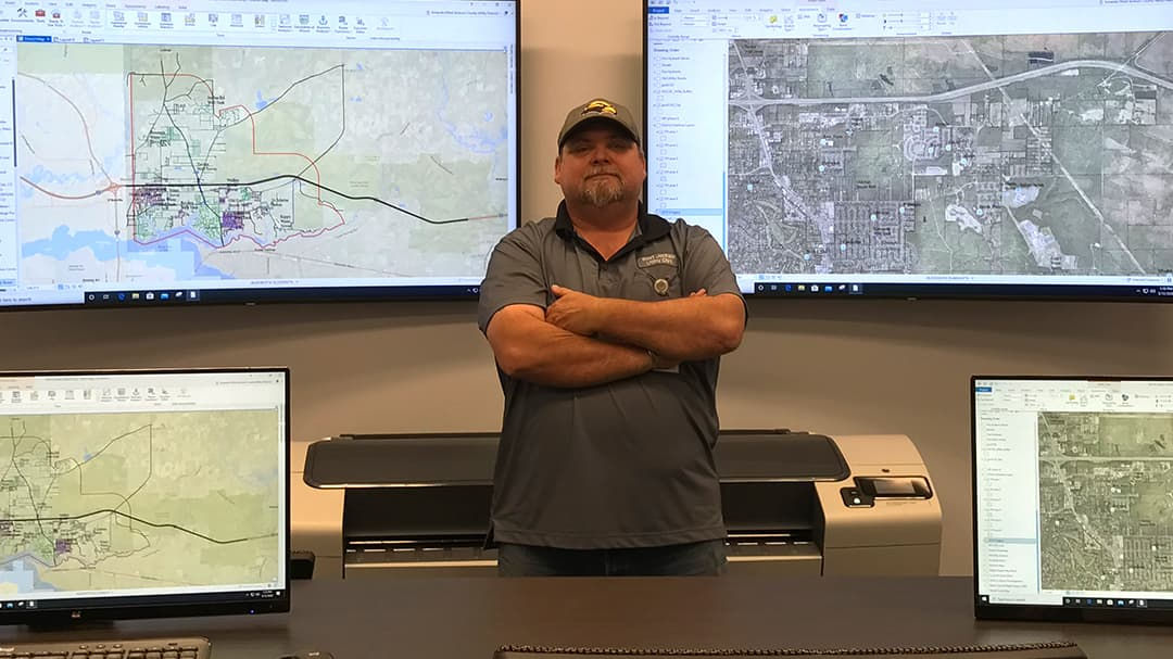 A person stands confidently in a GIS workspace surrounded by multiple screens displaying detailed maps and geographic data.
