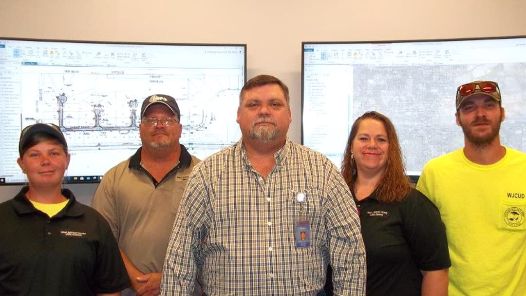 A group of individuals standing in an office environment with two large screens displaying technical diagrams and maps. 