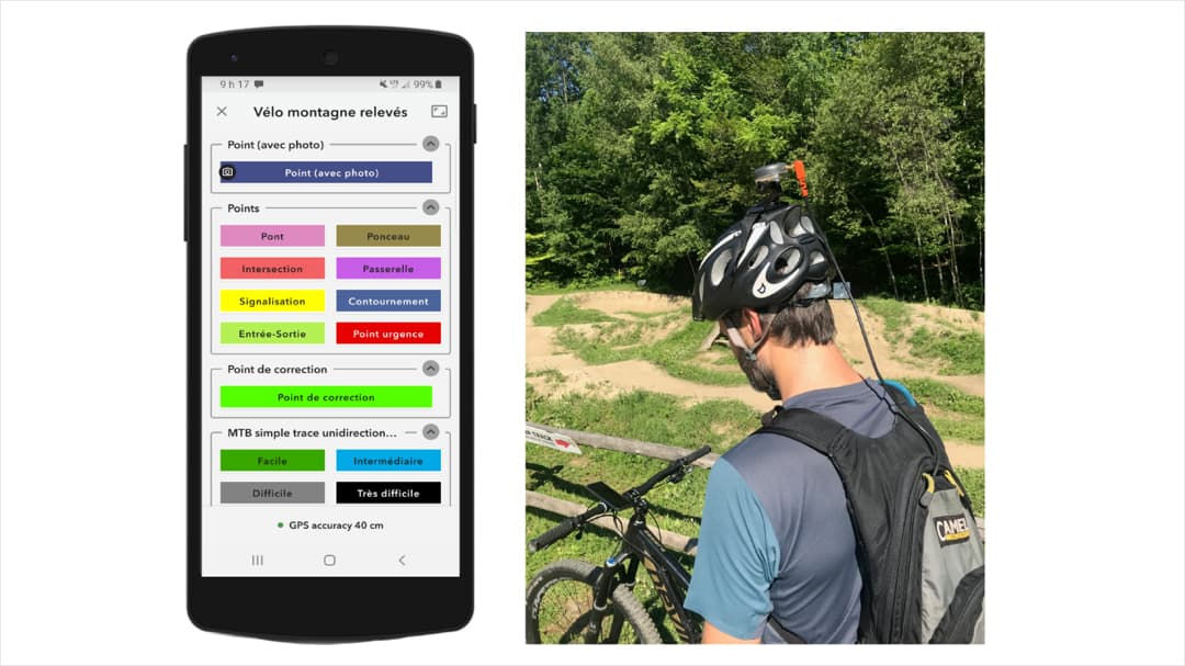 A man wearing a black helmet and backpack is seen using a smartphone app while standing near a dirt biking trail surrounded by greenery. 