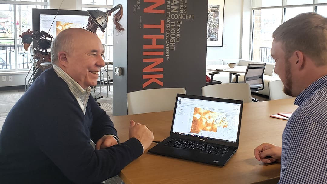 Two individuals are seated at a wooden table in a modern office setting, discussing content displayed on a laptop screen.