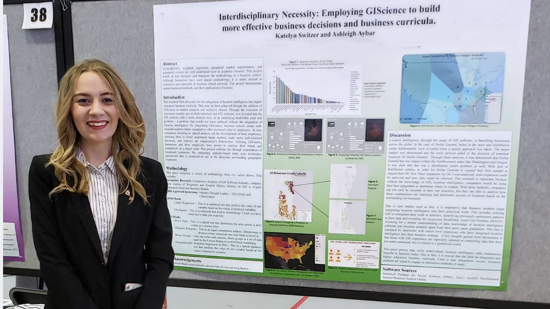 An academic poster titled 'Interdisciplinary Necessity: Employing GIScience to build more effective business decisions and business curricula' is displayed on a presentation board.