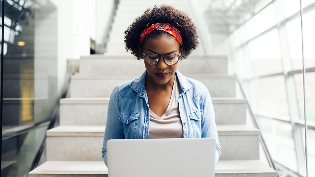 A woman sits on stairs in a bright, modern building while using a laptop. 