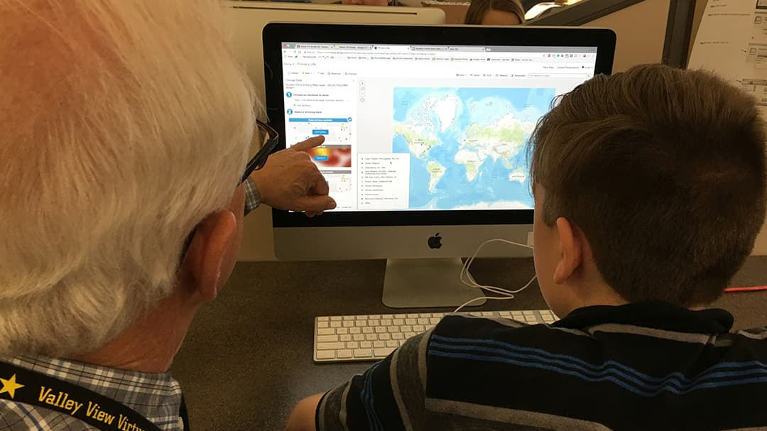 An older man and a young boy are seated at a desk, interacting with a desktop computer.