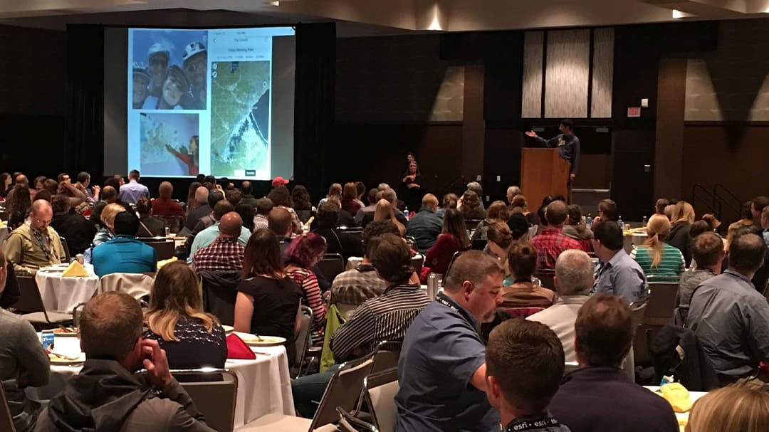 A large group of attendees seated in a conference hall, focusing on a presentation displayed on a screen. 