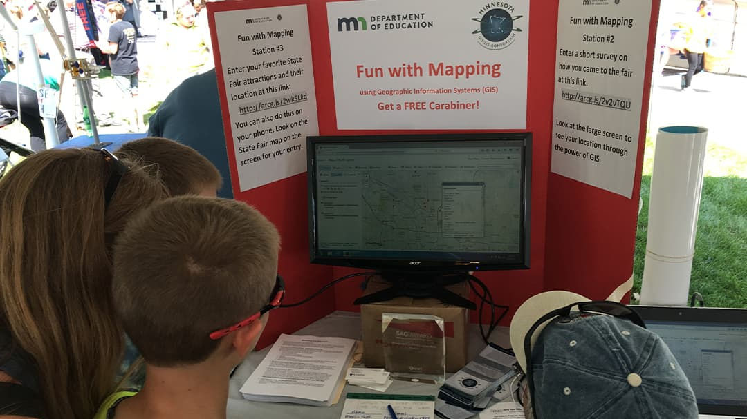 A group of children gathers around a computer screen displaying mapping software at an outdoor fair.