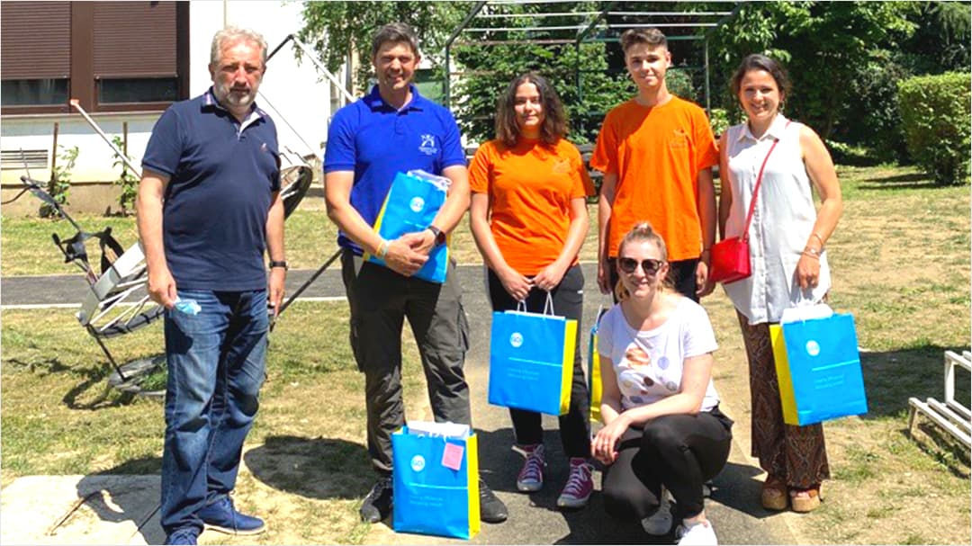 A group of people stands and kneels outdoors in a sunny setting, holding blue and yellow bags with visible circular designs.