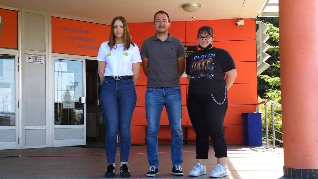 Three individuals are standing in front of a modern building with orange walls and glass doors.