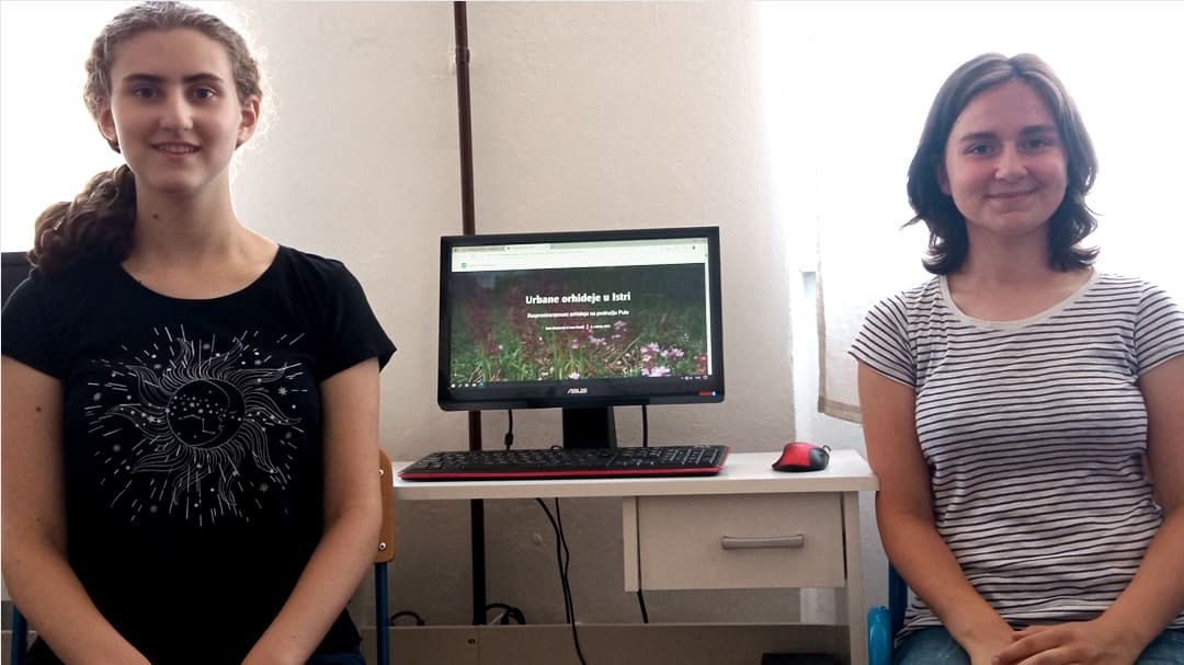 Two individuals are seated indoors near a desk with a computer monitor displaying a webpage featuring flowers and text. 