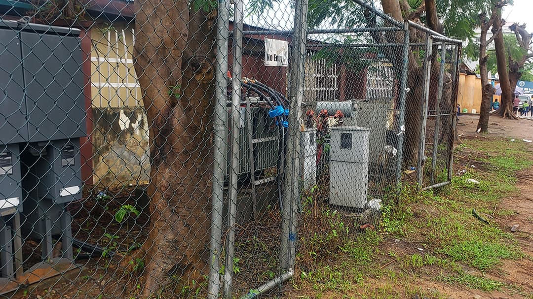 A fenced area containing various electrical equipment, including utility boxes and transformers, situated outdoors near trees and grass. 