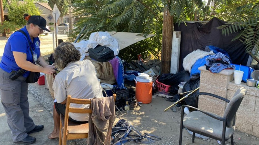 A person in a blue shirt provides aid to another seated individual near a makeshift shelter.