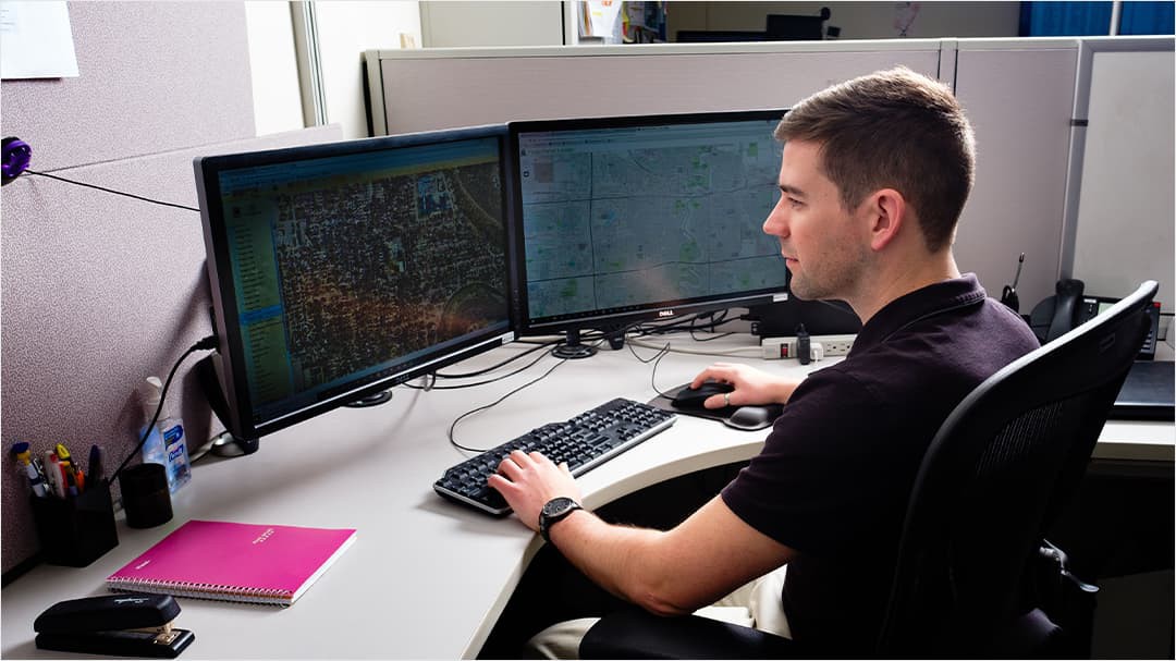 Man sitting at desk viewing two computer screens showing ArcGIS map and dashboard