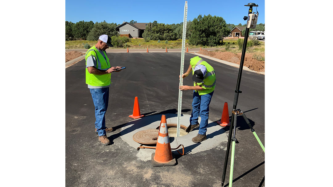 Two surveyors wearing high-visibility vests are inspecting a manhole on a newly paved road.