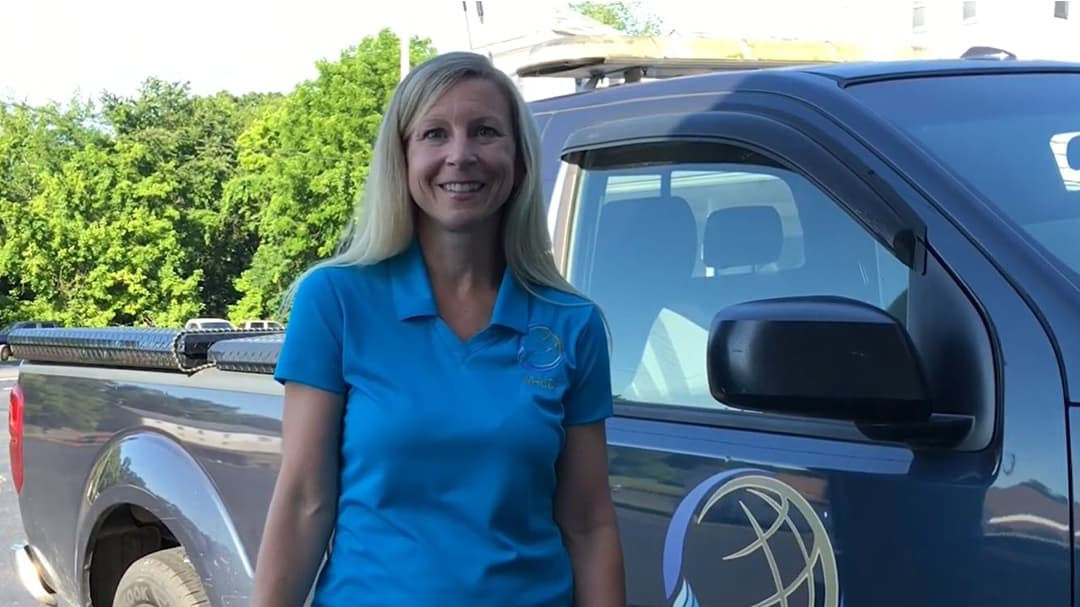 A woman wearing a blue polo shirt stands near a dark-colored truck with a visible globe graphic on the door. 