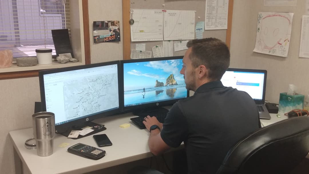 A person is seated at a desk working on a triple monitor setup in an office environment.