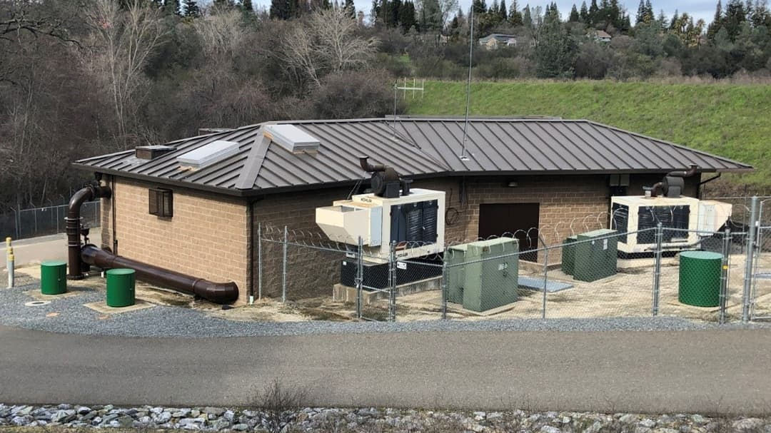 A small industrial building with a metal roof surrounded by fenced equipment, including large pipes and generators. 