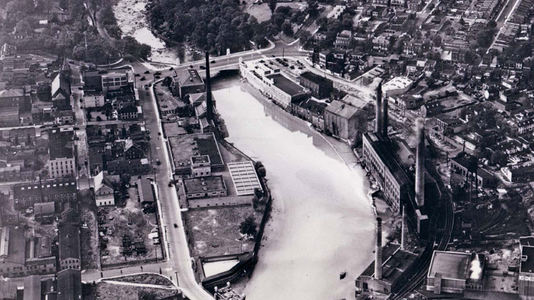 A black-and-white aerial photograph showcasing an industrial riverfront area with factories and smokestacks. 