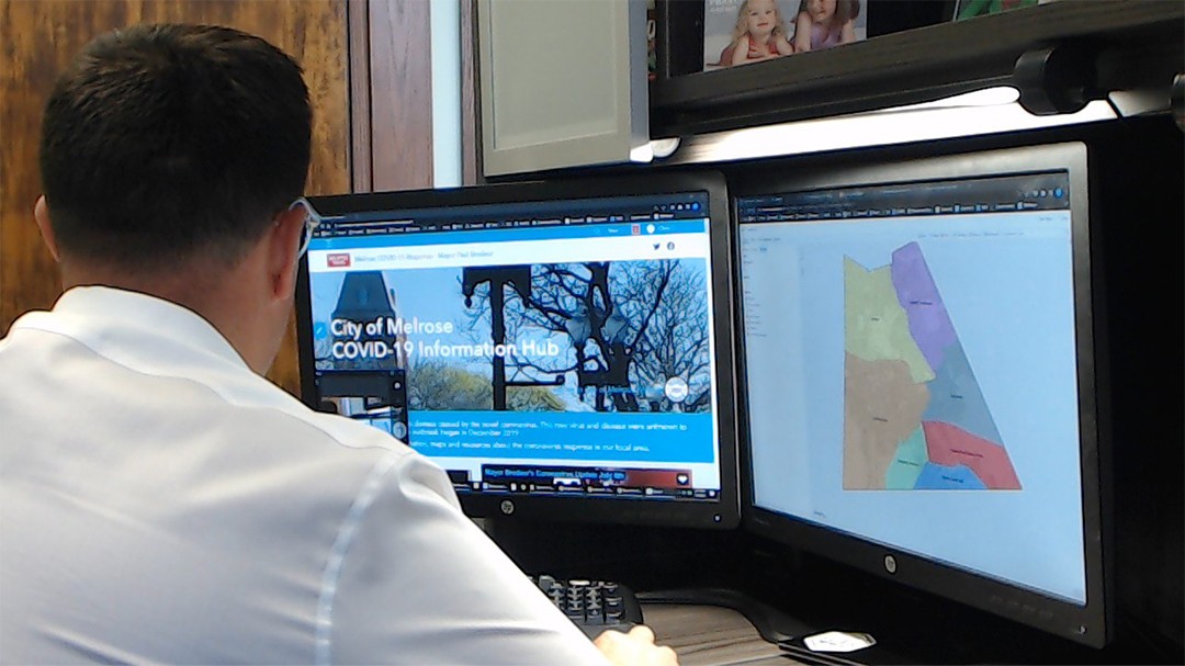 The back of a man's head who is facing two desktop computer monitor screens and looking at the homepage and a color coded map on the City of Melrose COVID-19 Information Hub