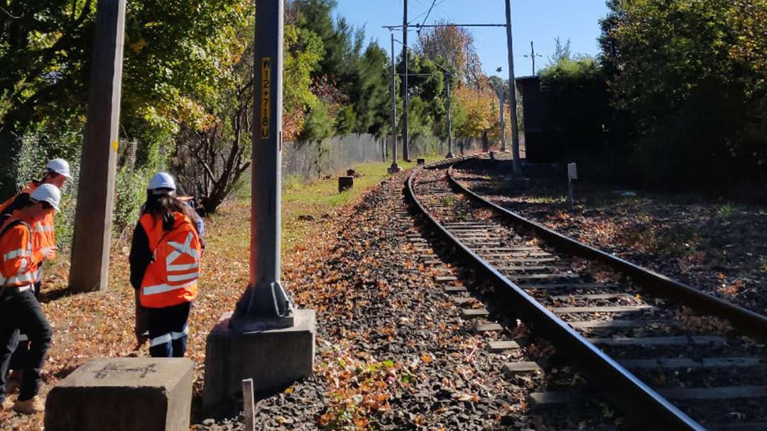 Two workers wearing high-visibility orange vests and helmets are inspecting railway tracks surrounded by autumn foliage. 