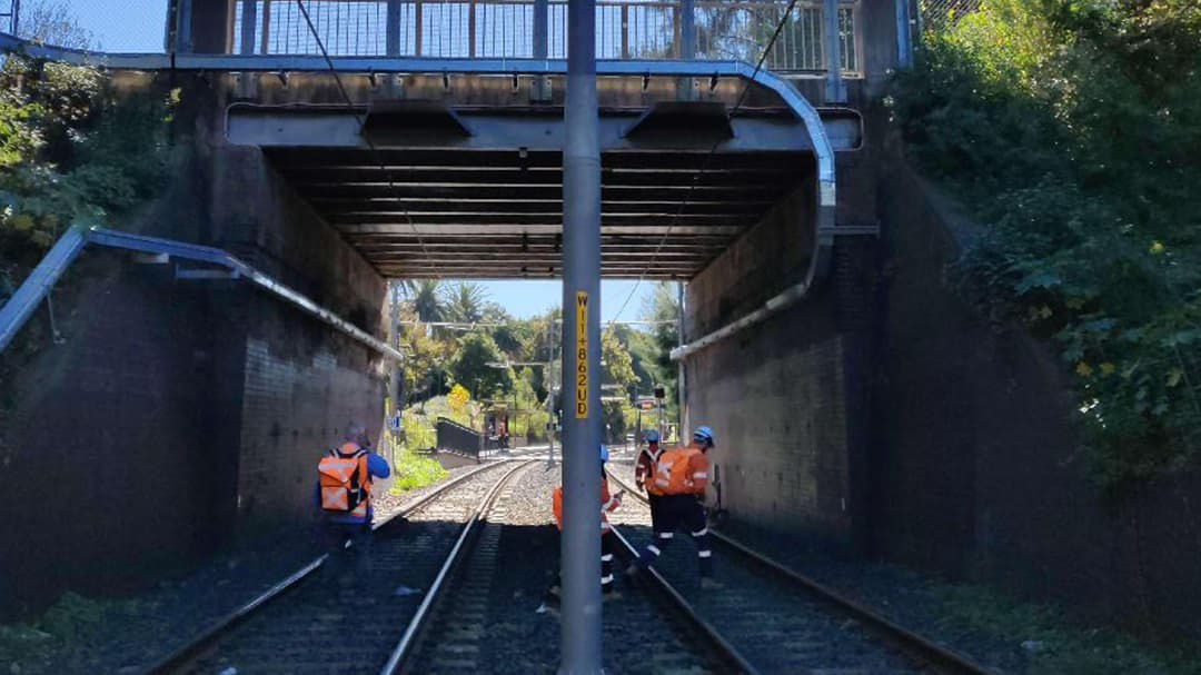 A group of workers in bright orange safety gear are seen on railway tracks beneath a bridge.