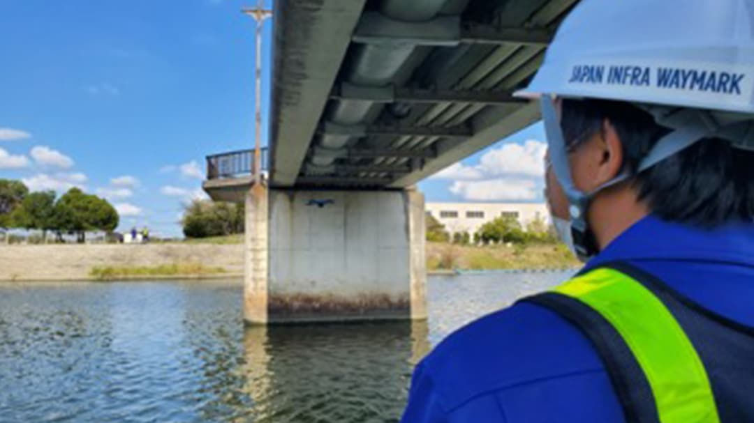 A person wearing a safety helmet and reflective vest is inspecting a bridge structure over a calm waterway. 