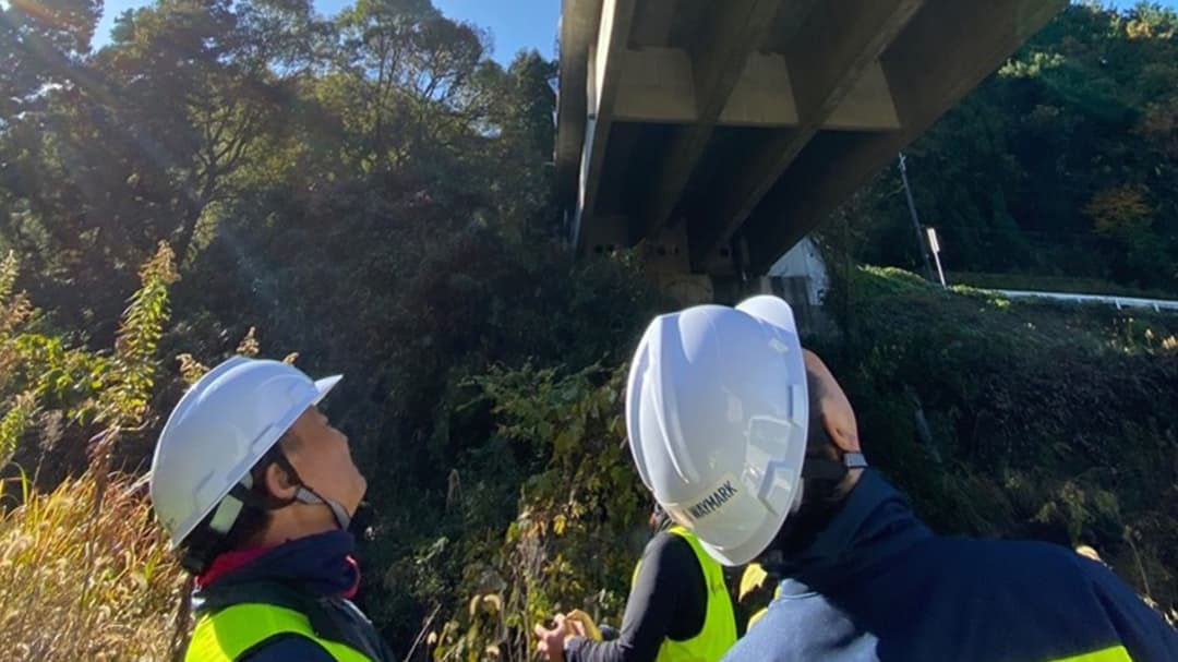 Two individuals wearing safety helmets and high-visibility vests are inspecting the underside of a bridge in a natural outdoor setting.
