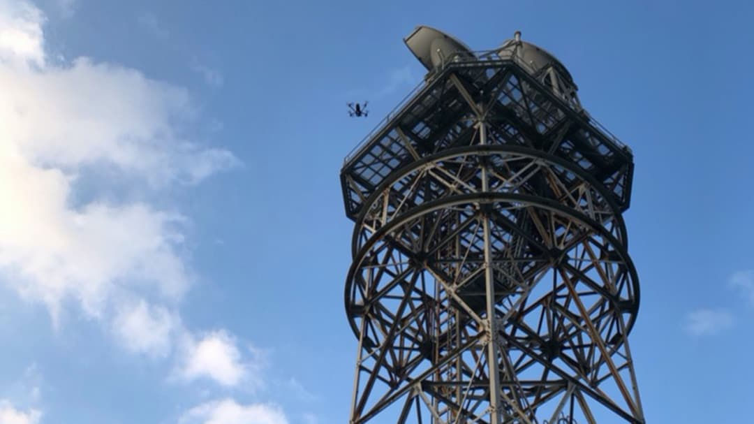 A drone is captured mid-flight near a tall, industrial-style metal tower against a backdrop of blue sky and scattered clouds.
