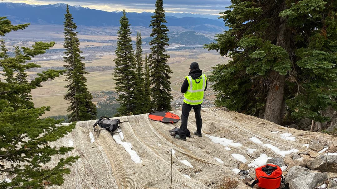 A person wearing a high-visibility vest stands on a rocky, snow-dusted mountain slope, preparing drone equipment.