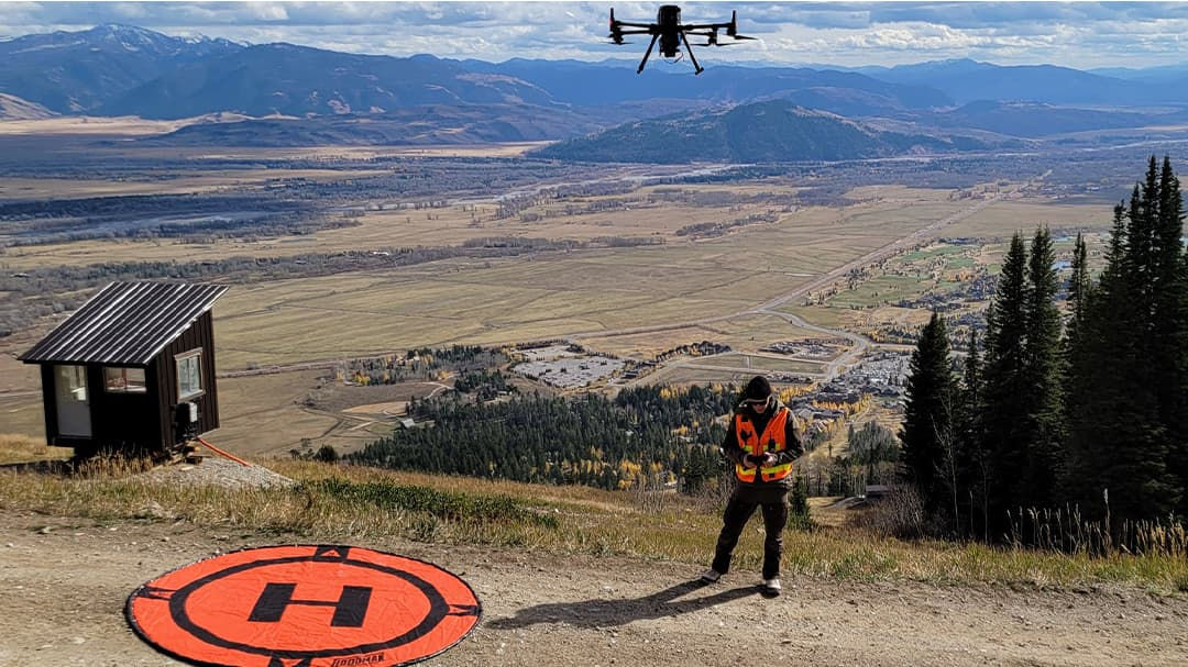 A drone operator wearing an orange safety vest stands near a marked landing pad in a mountainous area.