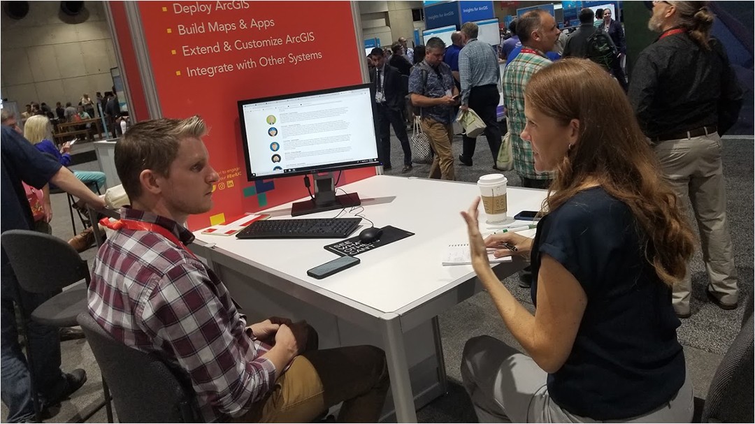 Karyn Nolan and Josh Peterson sit together at booth table during the 2019 Esri UC conference talking through her app ideas.