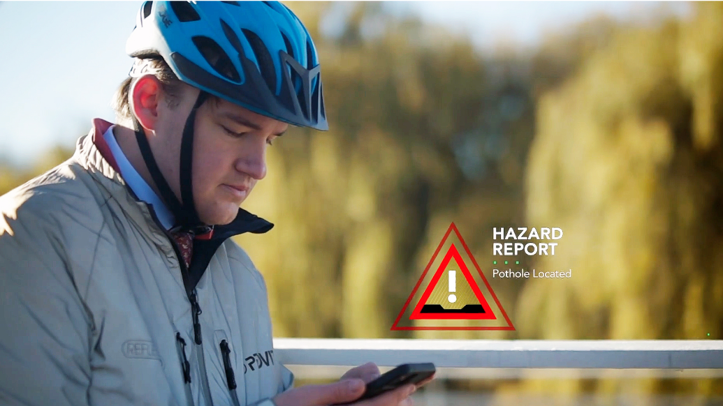 A person in a windbreaker and a bicycle helmet looking down at a mobile phone with sunlit trees in the background