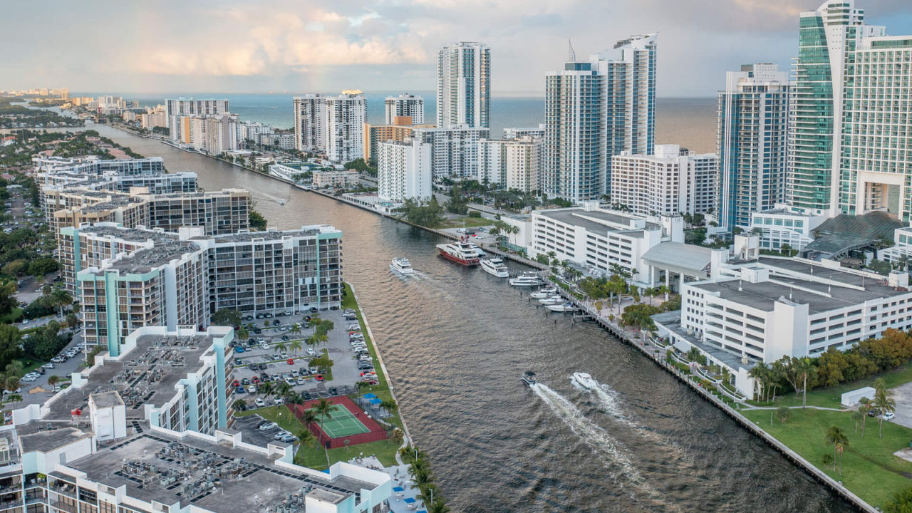 An aerial view captures a modern coastal city with high-rise residential and commercial buildings lining a wide canal. Several boats travel along the waterway, leaving visible wakes behind them. 