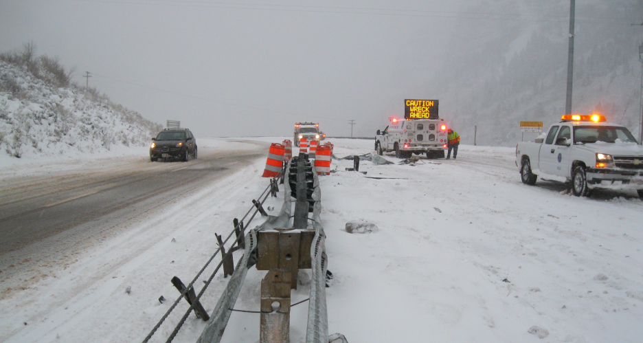 A snowy Utah highway shows a car near guardrails, safety barrels, and emergency trucks with signage warning of a wreck ahead