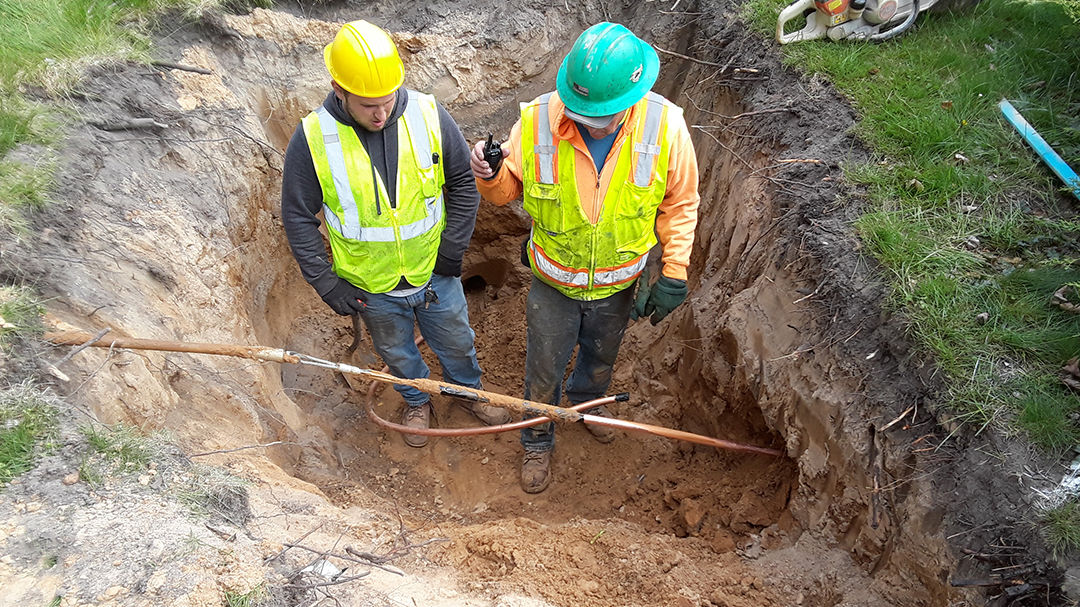 Two field staff in safety gear in a large hole in the ground performing a replacement. 